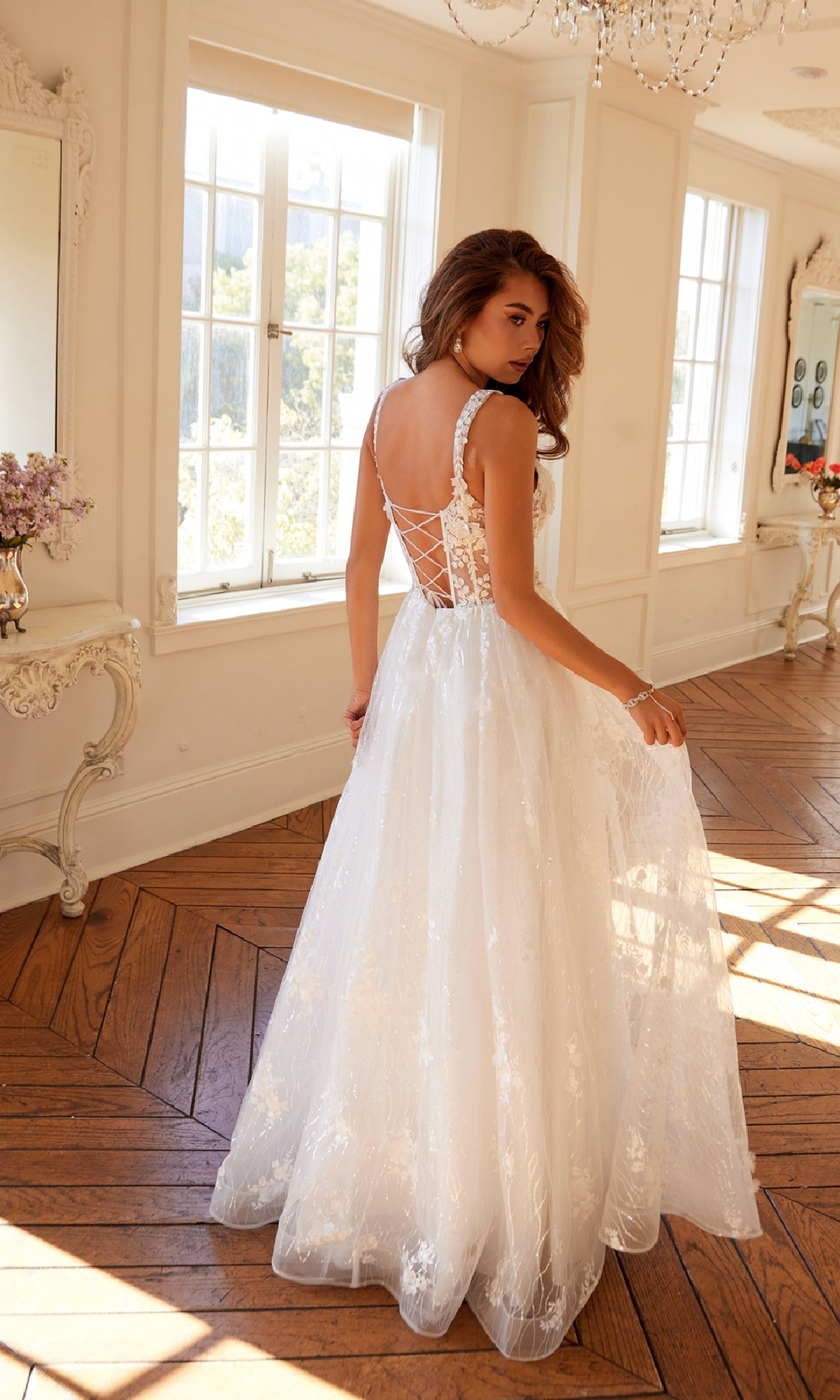 A woman wearing the Juliet JT2495AW long prom dress, featuring a white sleeveless lace corset bodice, stands in a bright room with wooden floors and large windows, looking over her shoulder.